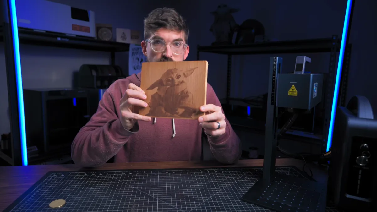 A presenter holds up a wooden workpiece engraved with a detailed image of Grogu, with a fiber laser on a post stand beside him.