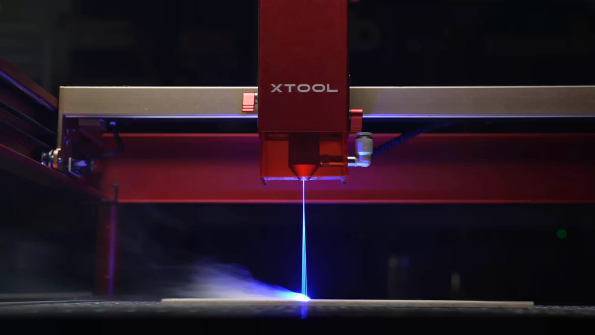 A diode laser head on a gantry with a visible blue beam running from the nozzle down to a thin board, throwing smoke and a blue glow across the work surface.