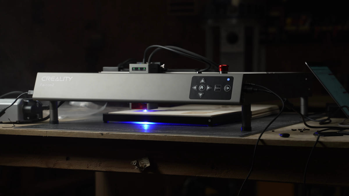 A Creality Falcon 2 diode laser on a workbench in a dark shop, engraving a workpiece with a bright blue point of light glowing under the laser head.