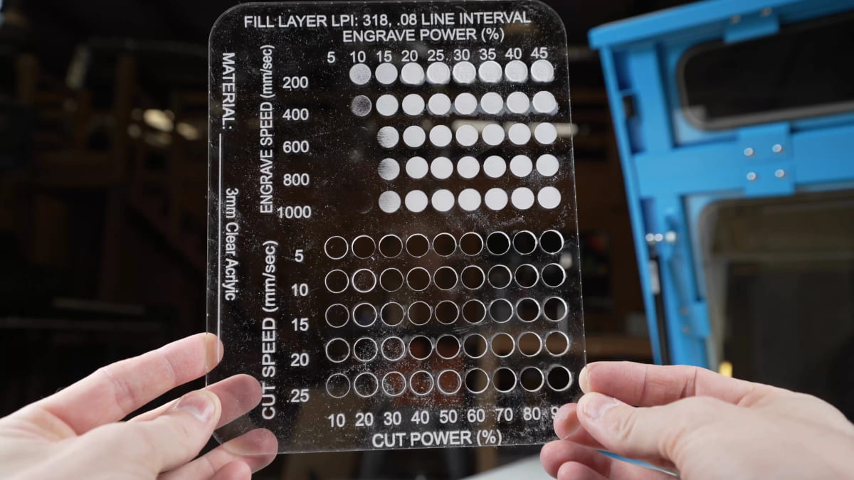 Two hands hold up a 3mm clear acrylic test grid, engraved and cut with a matrix of dots labelled by power and speed.