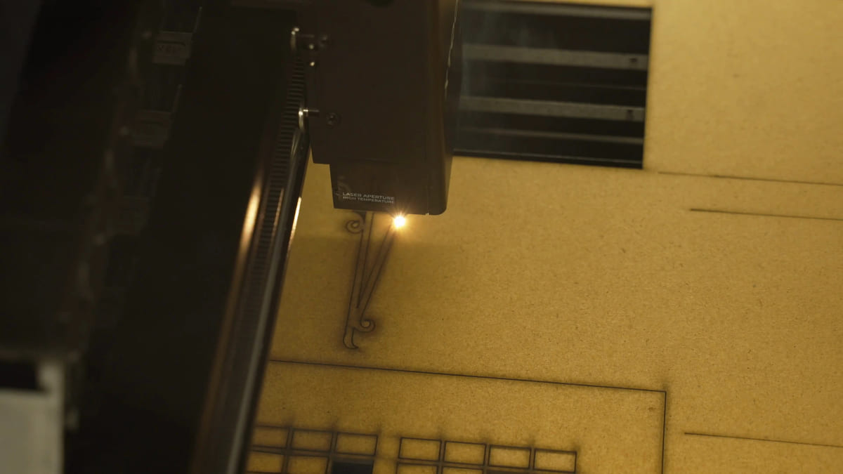 A laser head on its gantry cutting a curved line into a sheet of MDF, with a single bright orange point of light where the invisible beam meets the board.