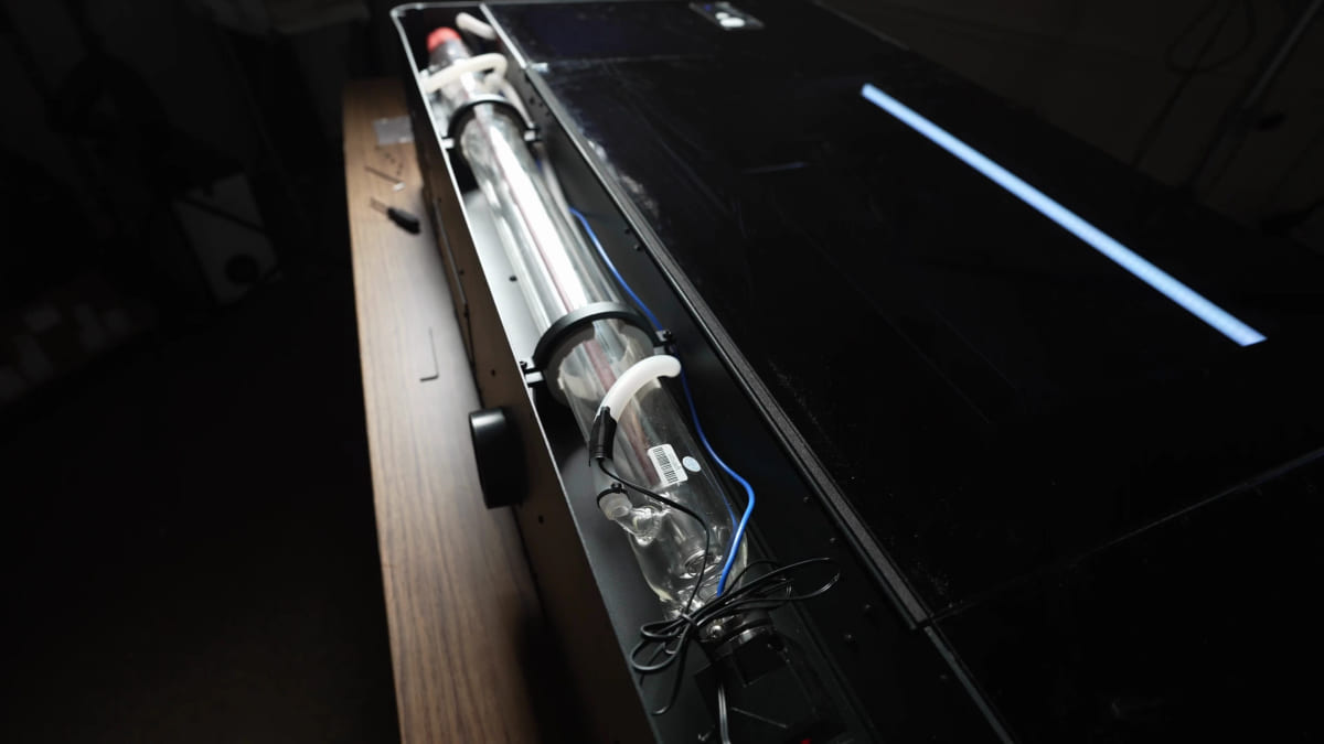 A long glass CO₂ laser tube with coolant fittings and wiring, lying in the rear compartment of a laser cutter.