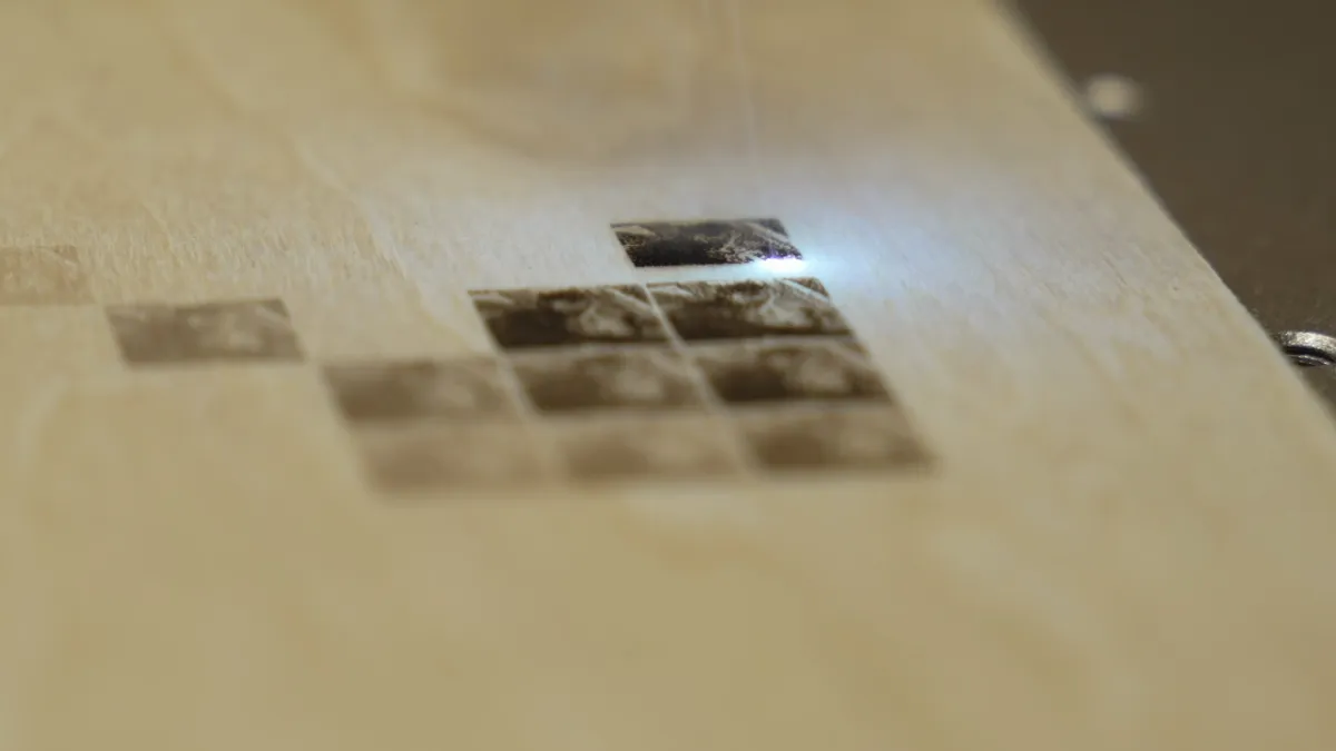 Macro view of pale birch with a block of engraved squares stepping from barely visible tan through to dark brown, the laser spot glowing bright on the square being engraved.