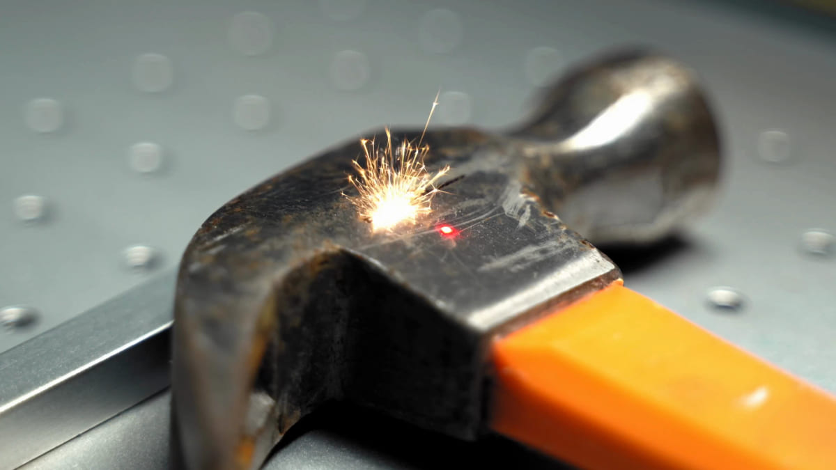 Close-up of a fiber laser mid-fire on the metal head of a claw hammer, a burst of sparks and a red aiming dot at the point where the beam lands.