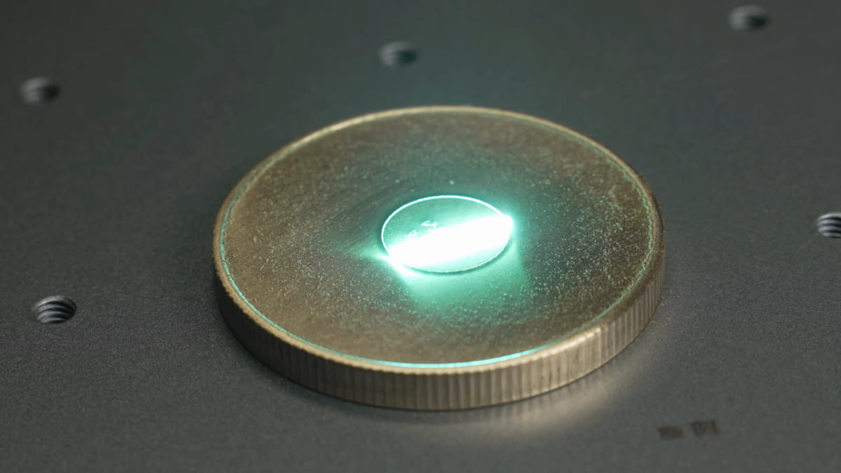 A brass-coloured coin lying on a dark machine bed with an intense blue-green bloom of light at the single point where a fiber laser is marking it.