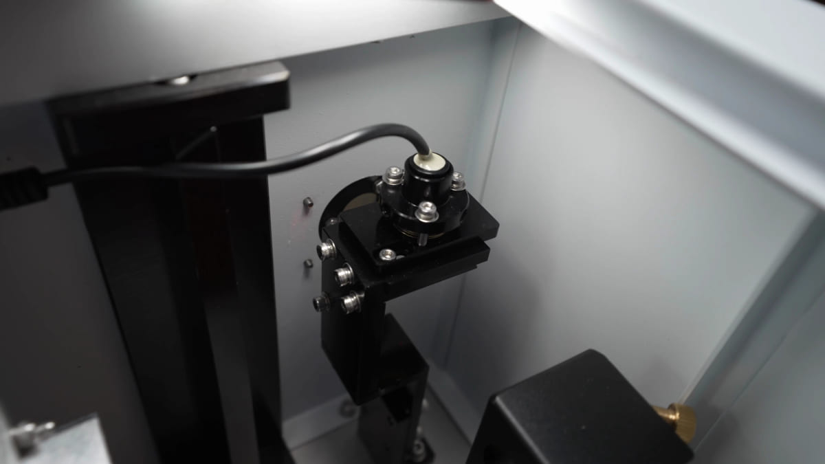 Close-up inside a laser machine of a round mirror held in a black bracket with adjustment screws, part of the beam path.
