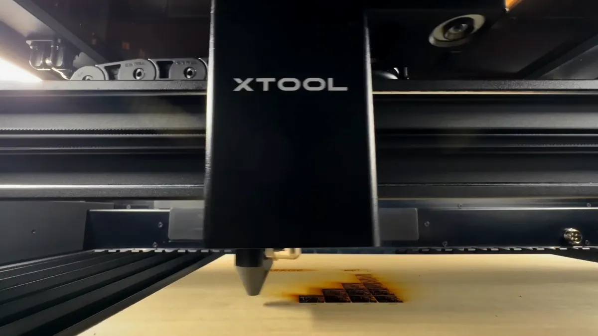 Close-up of a laser head travelling over a sheet of light wood, leaving a block of dark engraved squares behind it.