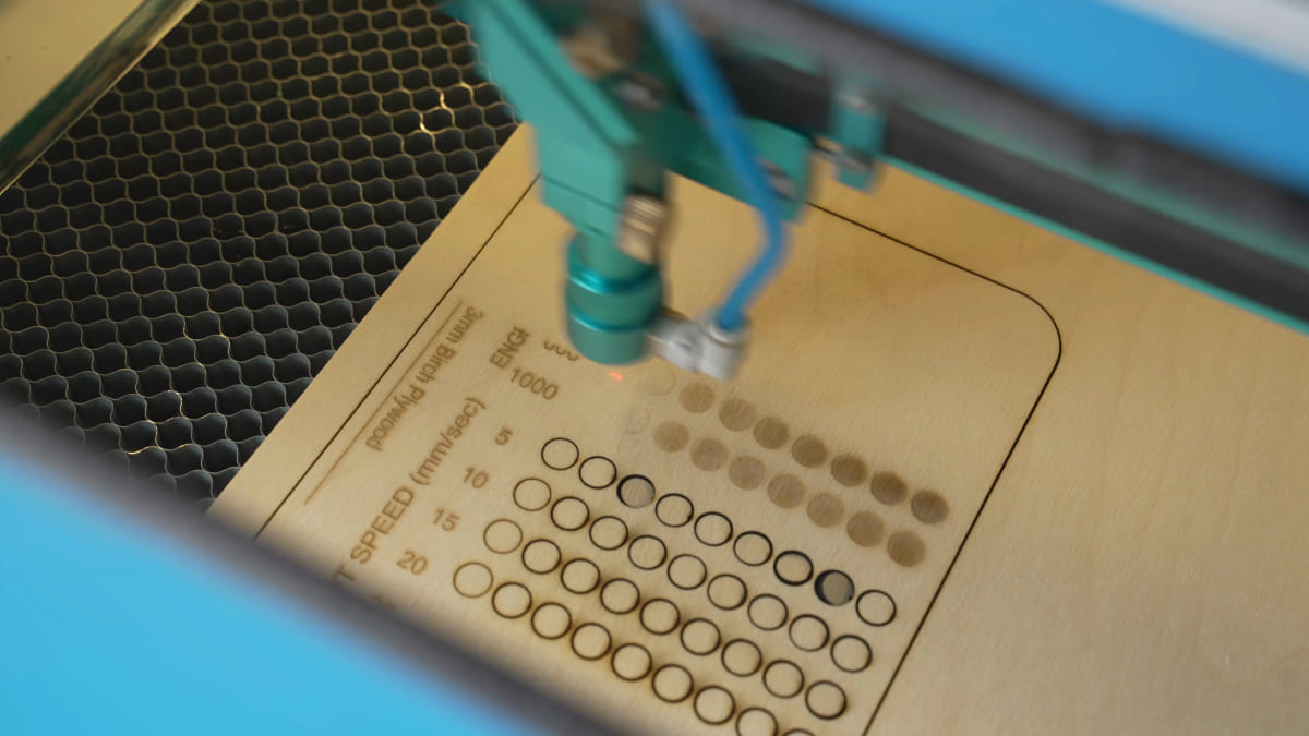 Close-up inside a laser cutter as the head engraves a grid of dots into 3mm birch plywood, with the rows labelled by speed in millimetres per second.