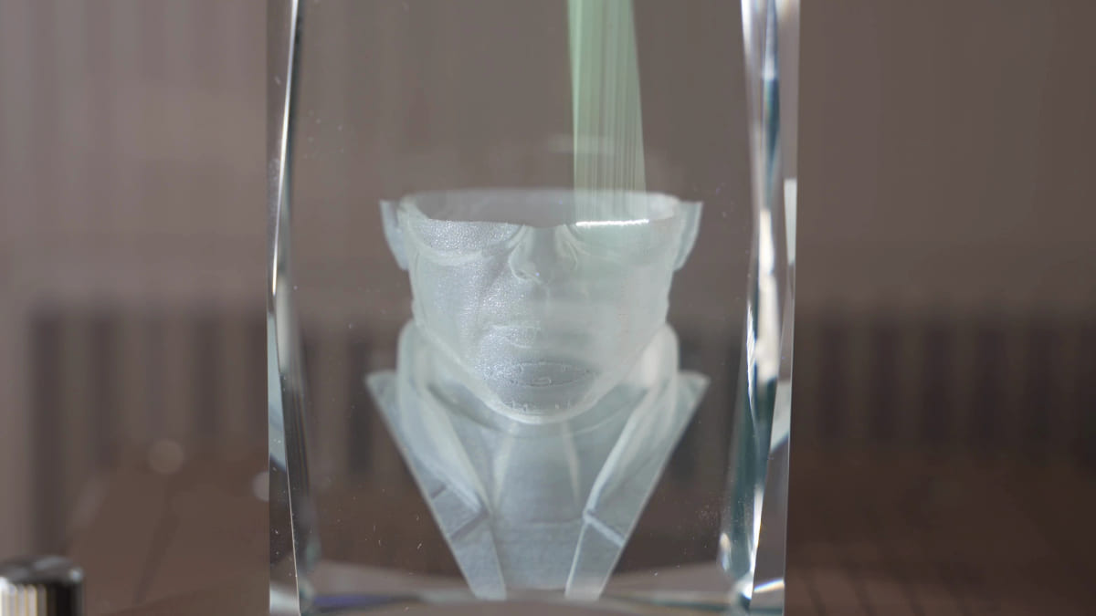 A clear glass block with a face being engraved inside it, the laser beam visible passing down through the glass to the frosted portrait forming in the middle.